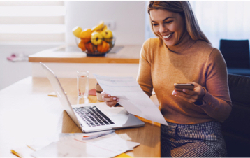 Een vrouw die met een laptop aan tafel zit, terwijl ze een mobiel en een vel papier vasthoudt.