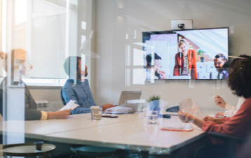 Three people sitting at a conference table looking at a mounted televesion screen with three people on the screen looking back.